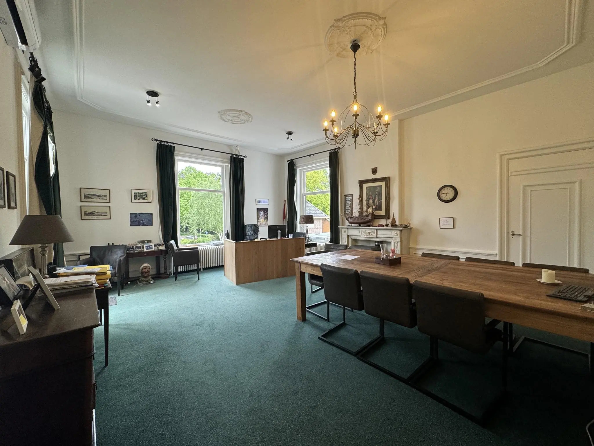 Ruim, klassiek ingericht kantoor aan de Dorpsstraat met houten vergadertafel, bureau bij grote ramen en groene vloerbedekking.